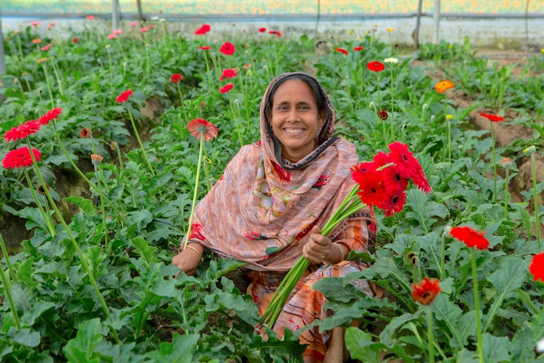 A woman in Bangladesh smiles as she holds red flowers in her greenhouse.