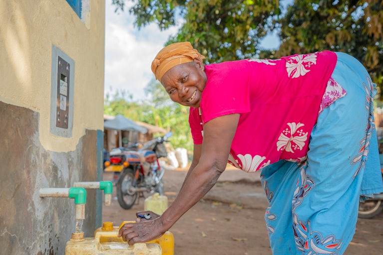 A woman smiling as she fills yellow jerry cans at a community water tap beside her home.