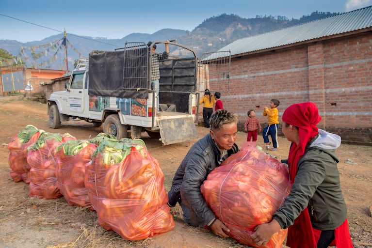 Two people lift a large bag of onions near several similar bags beside a white truck. Children play in the background, with mountains and brick buildings visible under a blue sky.