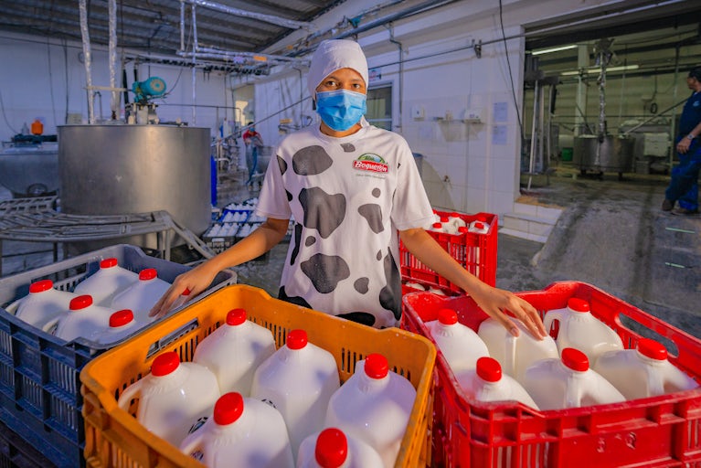 A woman wearing a blue medical mask standing in a factory behind two crates filled with gallon jugs of milk.