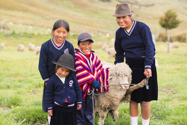 A group of four smiling children in sweaters and hats stands with a sheep on a grassy hillside. The scene is cheerful and pastoral.