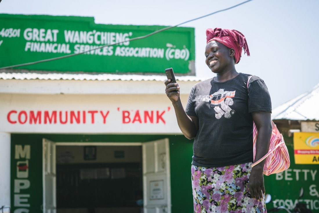 A Kenya woman, wearing a floral skirt, stands in front of a community bank and smiles at the cell phone she holds in her hand.