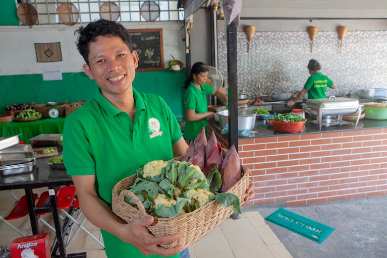 Smiling man in green shirt holds basket of fresh vegetables in commercial kitchen.