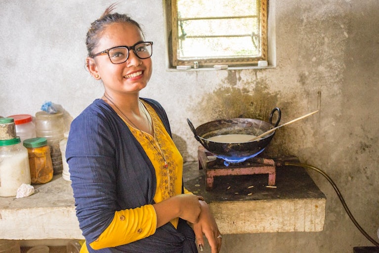 Smiling woman in glasses stands by cooking stove in rustic kitchen, wearing yellow and blue.