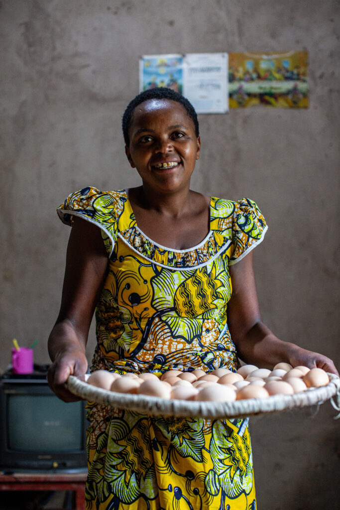 A woman in a yellow patterned dress holds a tray of eggs inside a chicken coop.