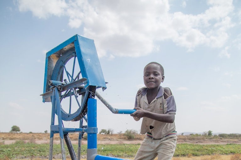 Boy operates water pump in arid agricultural setting.