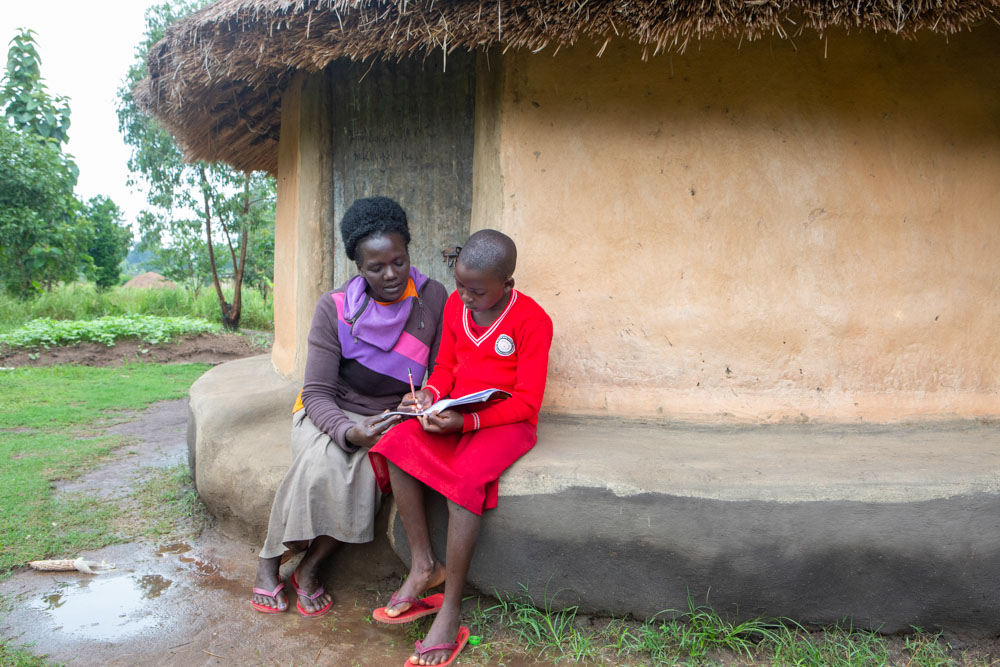 A woman seated beside a young girl wearing a school uniform shares a moment over a book outside their mud-walled home.