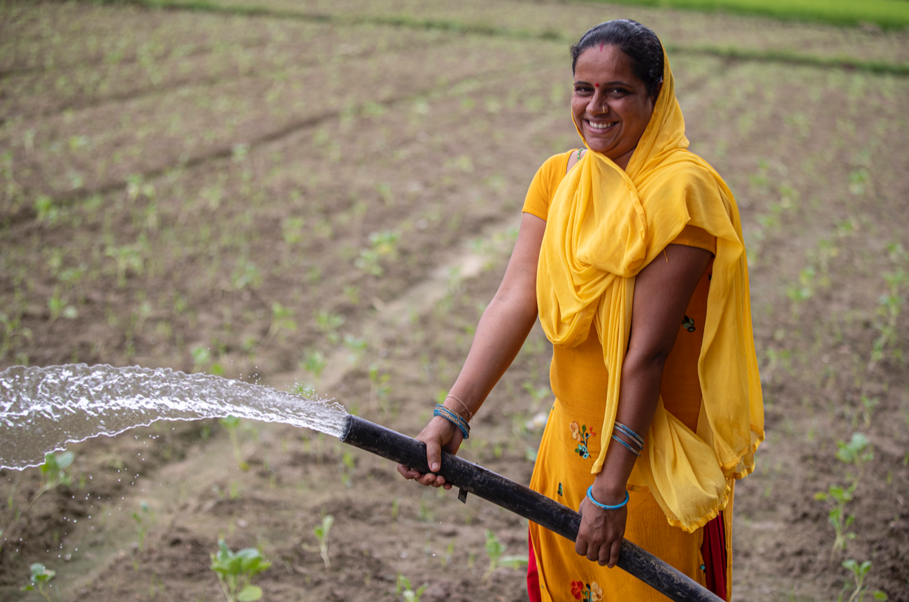 A woman wearing a yellow dress and scarf waters her field with a hose.